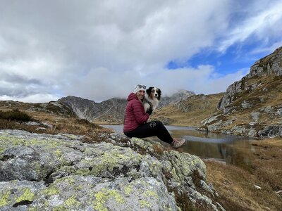 Person sitting on rocky outcrop with dog, surrounded by mountains and a lake, under an overcast sky with breaks of blue. | © Urlaub auf der Alm / Julia Winkler