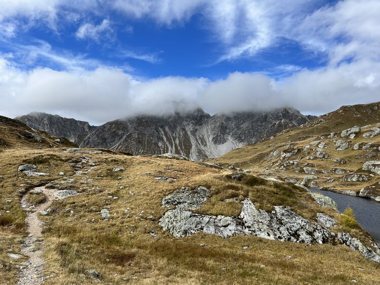 Rugged mountainous landscape with rocky terrain, grassy meadows, and low-hanging clouds obscuring the peaks. | © Urlaub auf der Alm / Julia Winkler