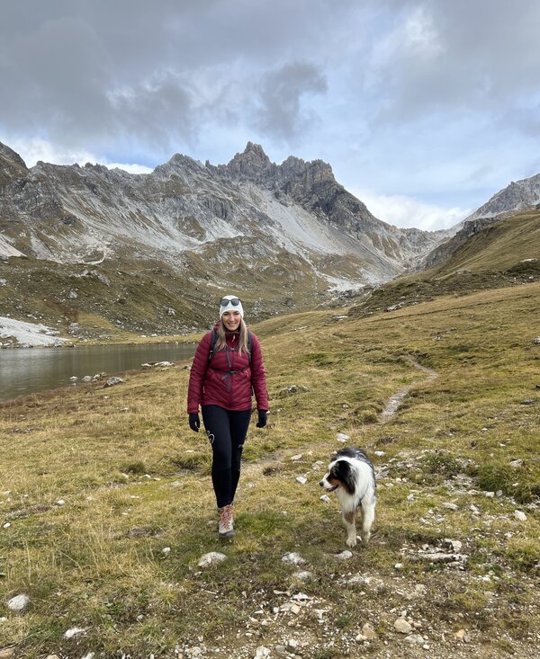 A woman in a red jacket walking with a dog in a mountainous, grassy landscape with snow-capped peaks in the background. | © Urlaub auf der Alm / Julia Winkler