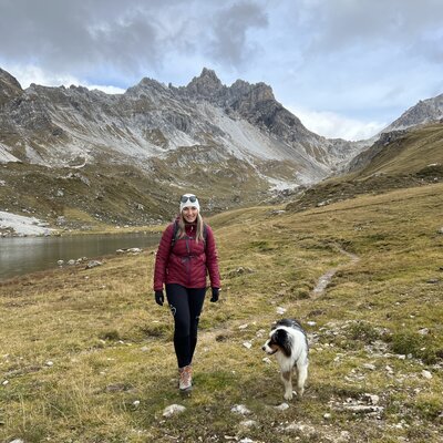 A woman in a red jacket walking with a dog in a mountainous, grassy landscape with snow-capped peaks in the background. | © Urlaub auf der Alm / Julia Winkler