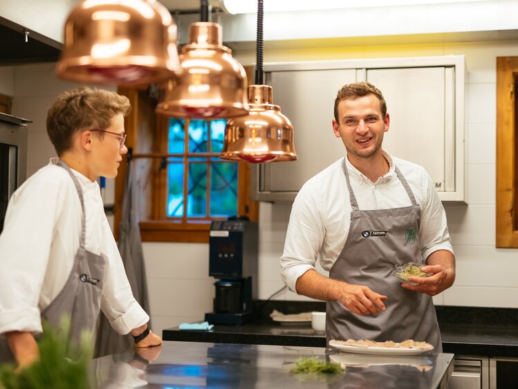 Two chefs in a kitchen, one preparing a dish while the other observes, surrounded by copper cookware and a window with a scenic view. | © Urlaub am Bauernhof / Daniel Gollner