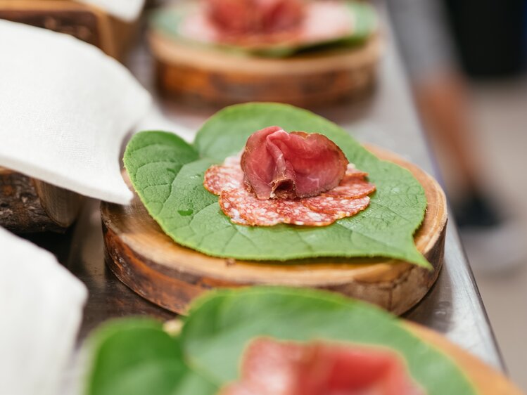 Wooden board with green leaves and slices of cured meat, creating an artistic food presentation. | © Urlaub am Bauernhof / Daniel Gollner