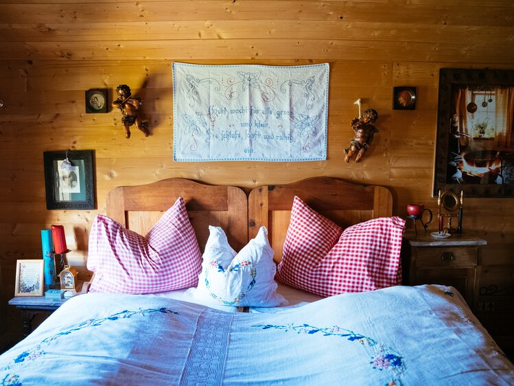 Rustic wooden cabin with embroidered textile artwork, antique decor, and cozy checked bedding. | © Urlaub am Bauernhof / Daniel Gollner