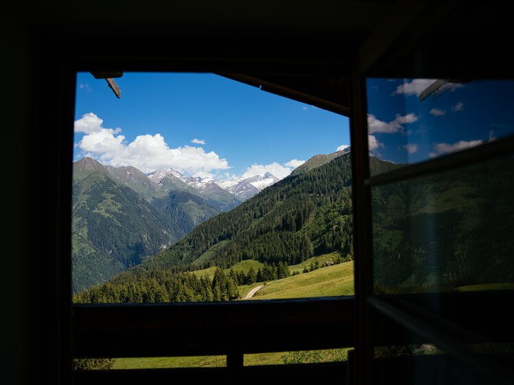 Scenic mountain landscape framed by wooden structure, lush forests, winding path, and distant snowy peaks against blue sky with clouds. | © Urlaub am Bauernhof / Daniel Gollner