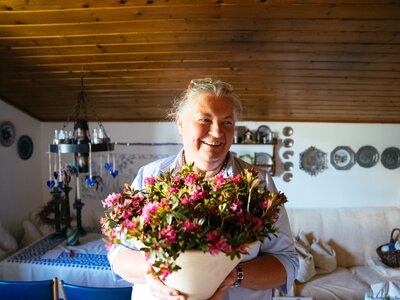 Gastgeberin Theresia Bacher mit einem Strauß Almrausch auf der Dürsteinalm | © Urlaub am Bauernhof / Daniel Gollner Smiling elderly woman holding a bouquet of colorful flowers in a cozy rustic interior. | © Urlaub am Bauernhof / Daniel Gollner