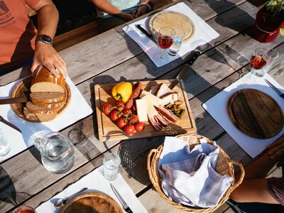 Rustic wooden table with fresh bread, cheese, tomatoes, and glasses of red wine. Outdoor setting with vibrant colors and textures. | © Urlaub am Bauernhof Steiermark / Daniel Gollner