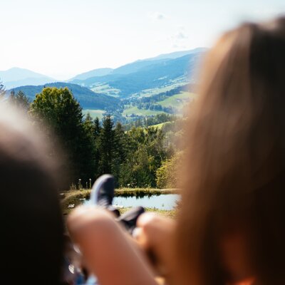 A tranquil mountain landscape with a person relaxing in the foreground, surrounded by lush forests, a lake, and distant peaks. | © Urlaub am Bauernhof Steiermark / Daniel Gollner