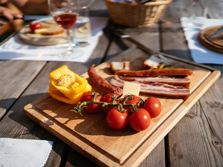Wooden cutting board with fresh tomatoes, bacon, and a pepper slice on a rustic table setting with wine glass. | © Urlaub am Bauernhof Steiermark / Daniel Gollner 