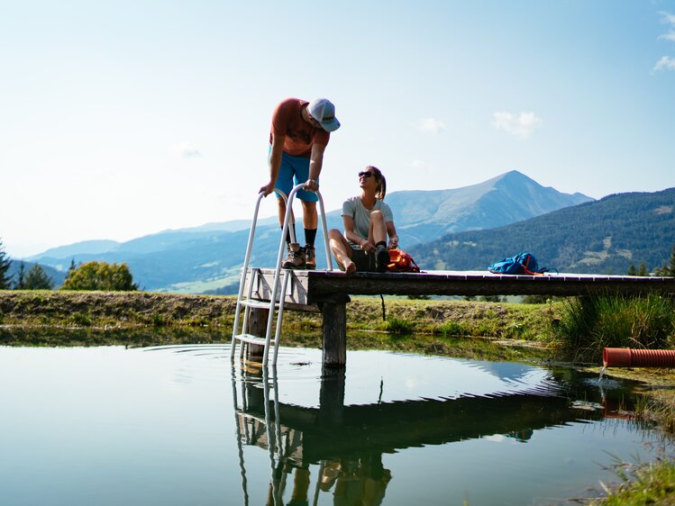 Two people sitting on a wooden deck overlooking a tranquil lake surrounded by mountains. | © Urlaub am Bauernhof Steiermark / Daniel Gollner 