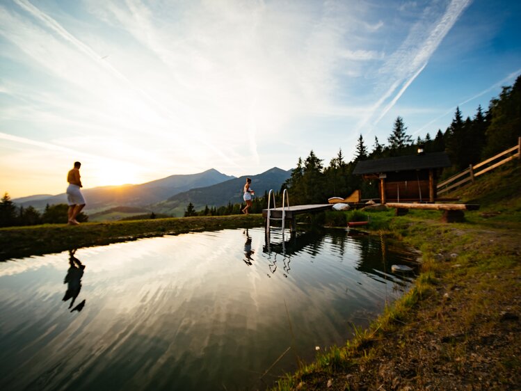 Picturesque mountain landscape with a small pool, wooden dock, and silhouettes of people at sunset. | © Urlaub am Bauernhof Steiermark / Daniel Gollner 