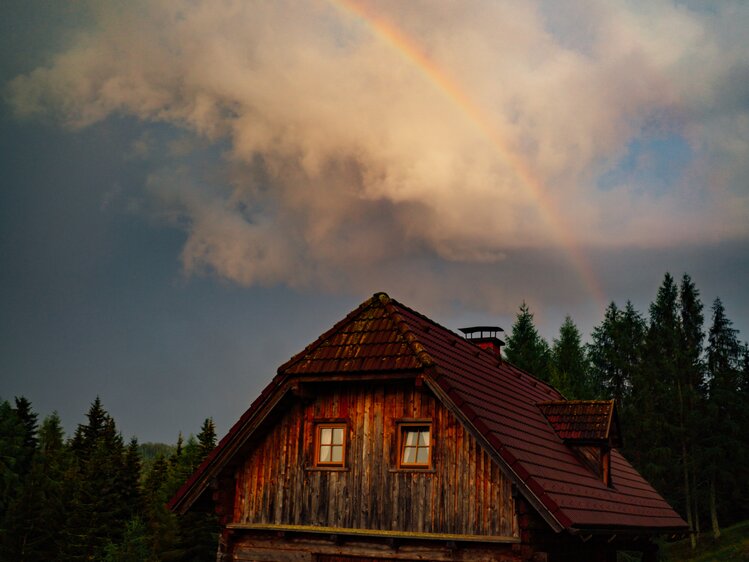 Rustic wooden cabin surrounded by pine trees, with a vibrant rainbow in the cloudy sky above. | © Urlaub am Bauernhof Steiermark / Daniel Gollner 