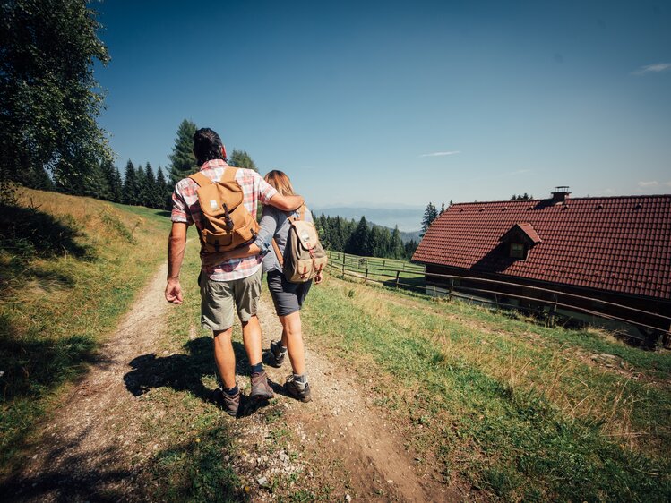 Two hikers with backpacks walking on a rural path with a traditional cabin in the background, surrounded by a scenic mountainous landscape. | © Urlaub am Bauernhof Steiermark / Daniel Gollner 