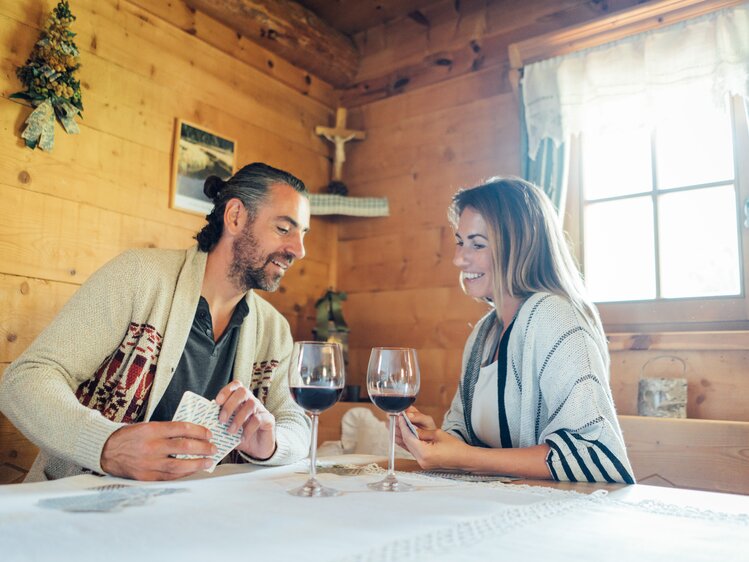 A smiling couple enjoying wine and conversation in a cozy, wooden cabin setting with a crucifix and Christmas decorations. | © Urlaub am Bauernhof Steiermark / Daniel Gollner 
