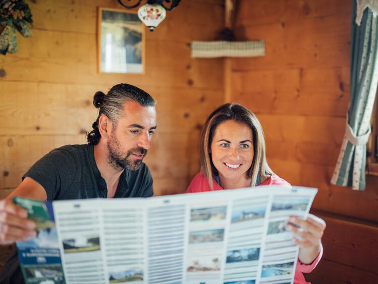 Couple in a rustic cabin, smiling and looking at travel brochures, cozy and relaxed atmosphere. | © Urlaub am Bauernhof Steiermark / Daniel Gollner 
