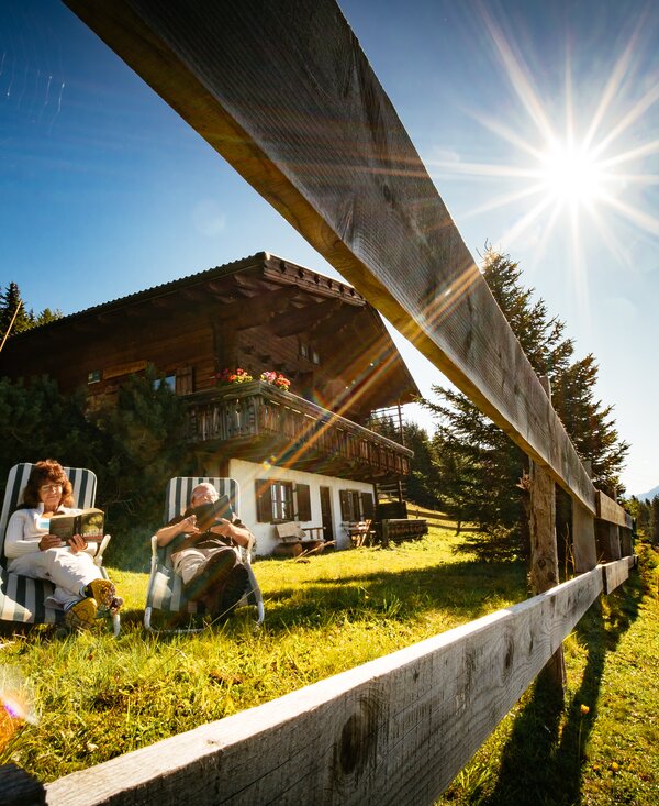 Wooden chalet surrounded by lush greenery, with two people relaxing in chairs under the warm sun's rays, against a backdrop of forested mountains. | © Urlaub am Bauernhof Tirol / Daniel Gollner 