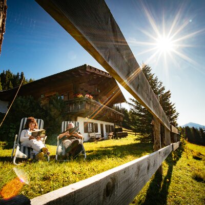 Wooden chalet surrounded by lush greenery, with two people relaxing in chairs under the warm sun's rays, against a backdrop of forested mountains. | © Urlaub am Bauernhof Tirol / Daniel Gollner 