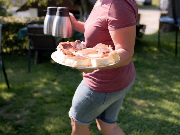 Person holding a plate of grilled meat and cheese in a garden on a sunny day.