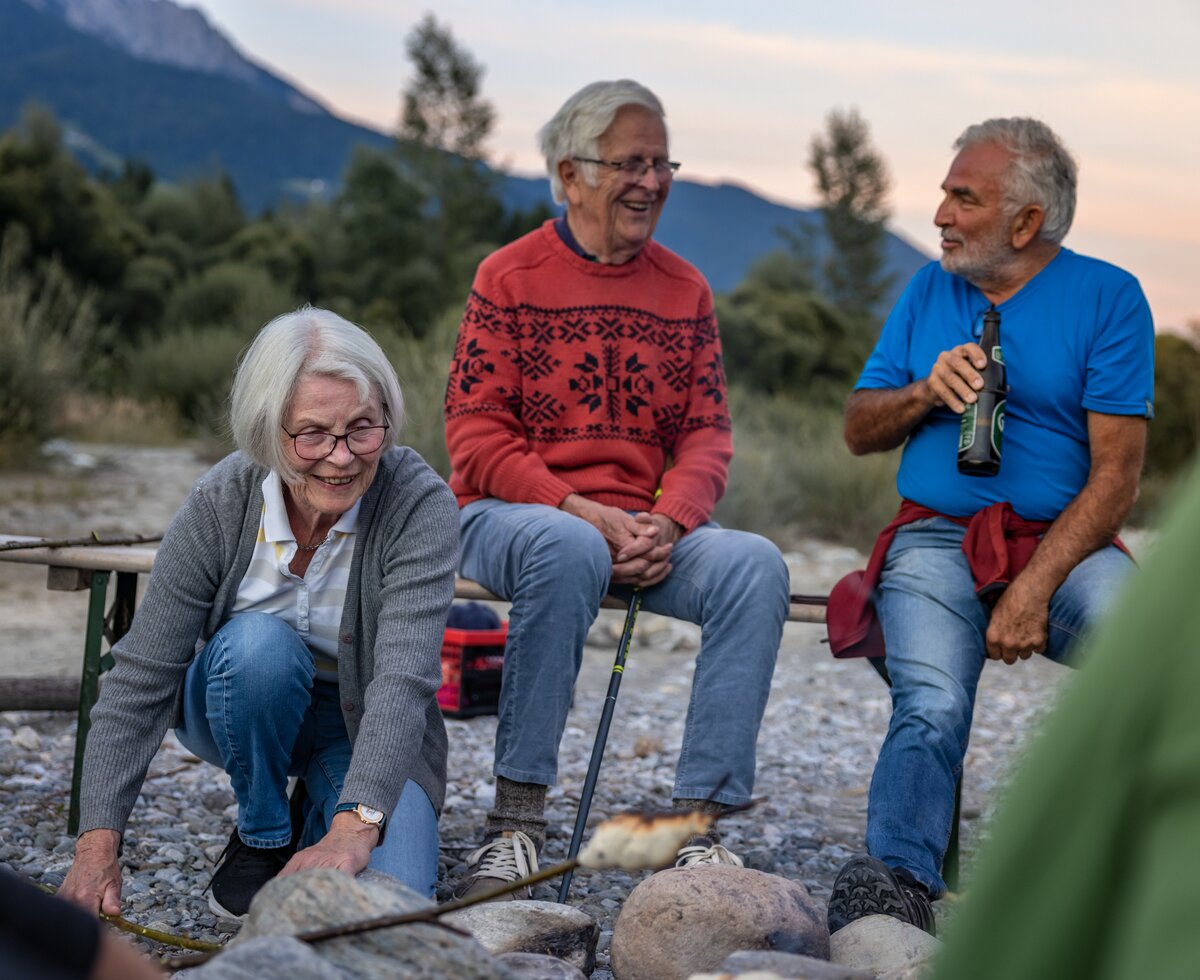Familie sitzt am Lagerfeuer | © Landesverband Urlaub am Bauernhof Kärnten/ Achim Mandler Männer unterhalten sich und die Frau grillt das Stockbrot im Lagerfeuer | © Landesverband Urlaub am Bauernhof Kärnten/ Achim Mandler