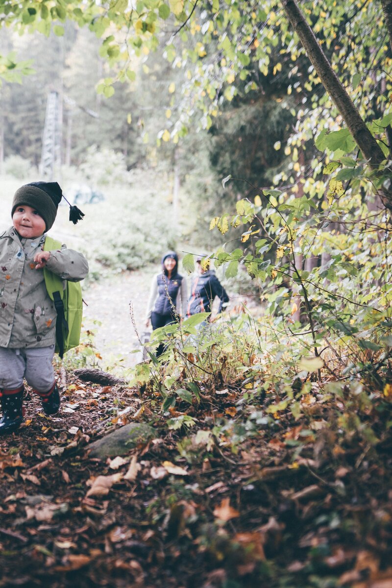 A small child in casual autumn attire exploring a lush, wooded path, with two adults walking behind in the distance. | © Urlaub am Bauernhof / Daniel Gollner