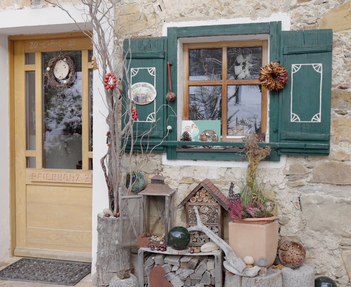 Picturesque cottage entrance with rustic stone walls, decorated with wreaths, shutters, and a cozy arrangement of potted plants and ornaments. | © Urlaub am Bauernhof / Daniel Gollner