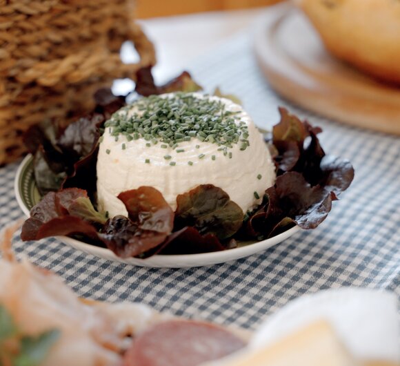 Cream cheese spread garnished with green herbs, served on a plate with lettuce leaves, accompanied by a wicker basket and a baked item. | © Urlaub am Bauernhof / Daniel Gollner