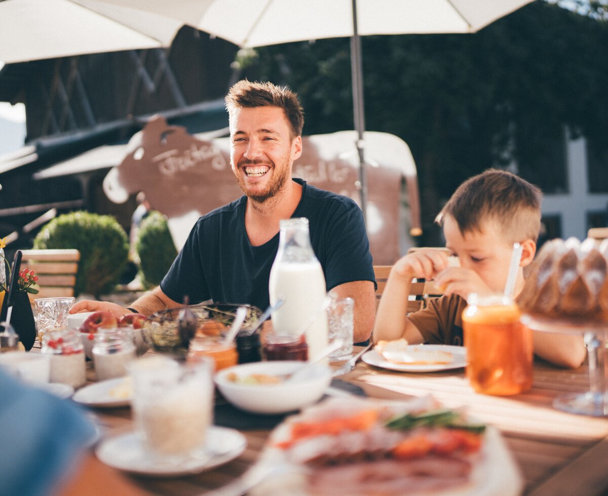 Outdoor cafe setting with smiling man, child, drinks, and food on wooden table under market umbrellas. | © Urlaub am Bauernhof / Daniel Gollner