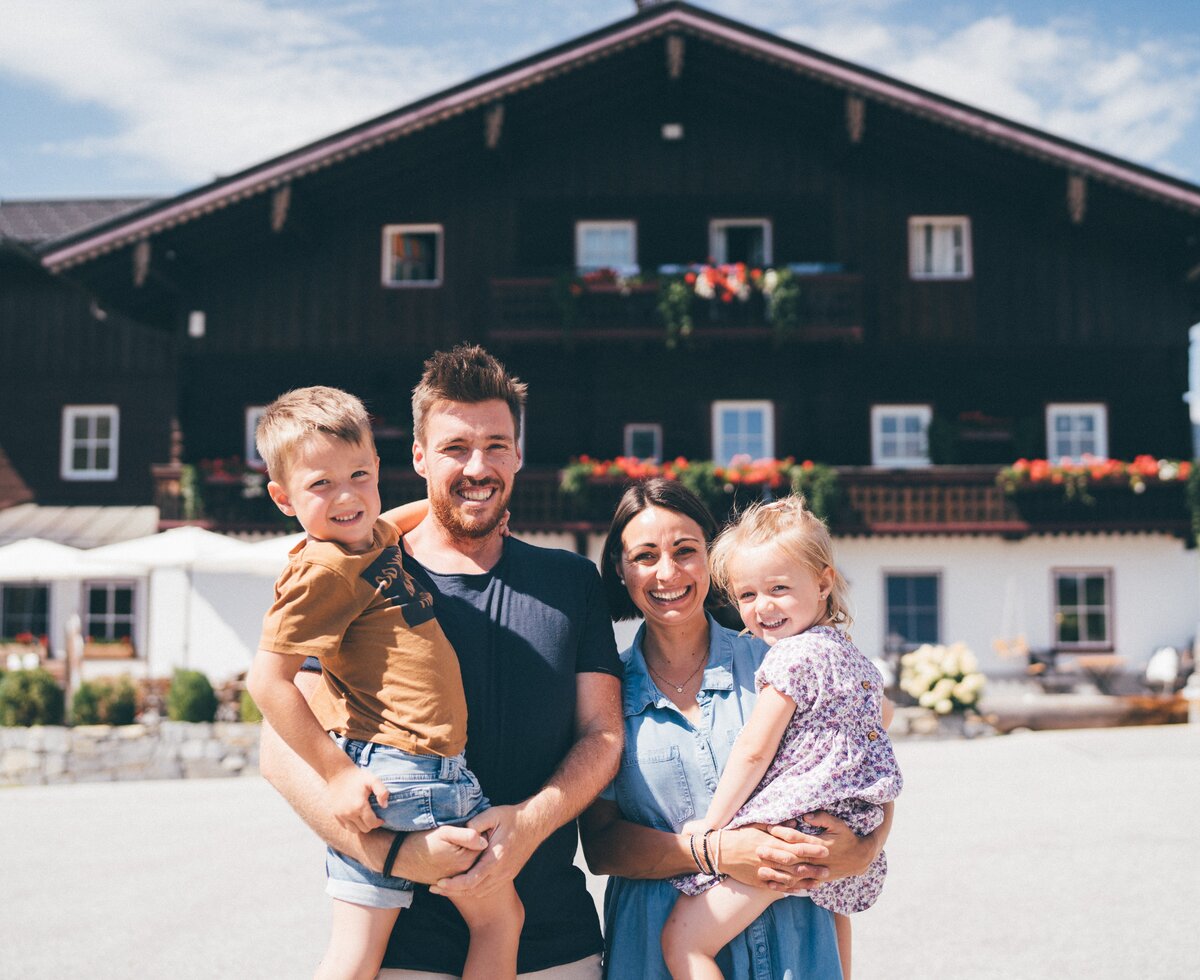 Happy family posing in front of a traditional wooden house with flowers and greenery. | © Urlaub am Bauernhof / Daniel Gollner