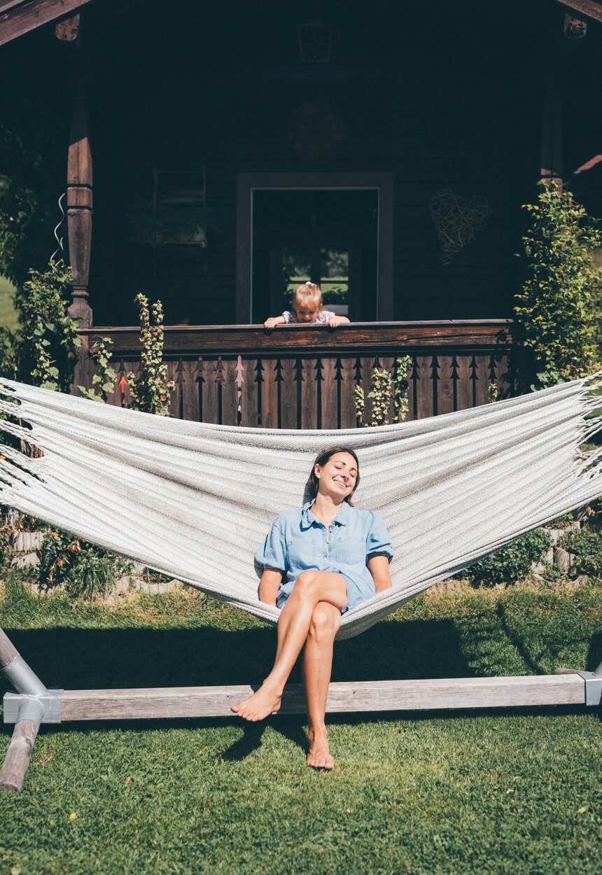 Young smiling woman relaxing in hammock on wooden porch, surrounded by lush greenery and rustic cabin. | © Urlaub am Bauernhof / Daniel Gollner