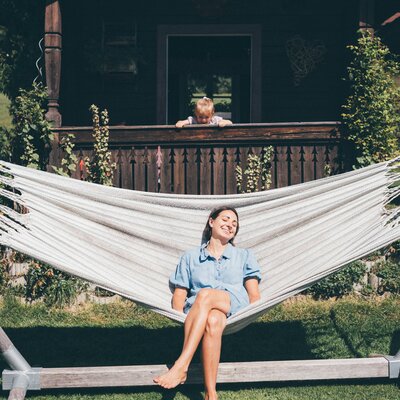 Young smiling woman relaxing in hammock on wooden porch, surrounded by lush greenery and rustic cabin. | © Urlaub am Bauernhof / Daniel Gollner