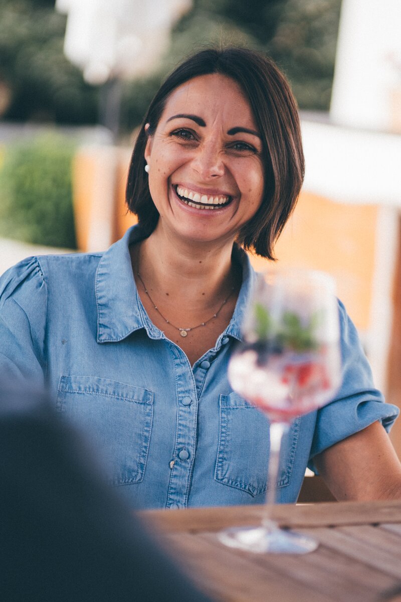 Woman sitting outdoors at a cafe table, smiling and drinking from a glass. | © Urlaub am Bauernhof / Daniel Gollner