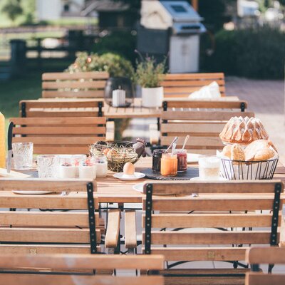Cozy outdoor dining setting with wooden benches, table, fresh pastries, juice, and decorative elements on a sunny day. | © Urlaub am Bauernhof / Daniel Gollner