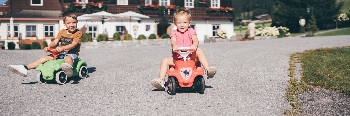 Two young children riding toy cars in a charming rural setting, with a traditional alpine-style building in the background adorned with flower boxes. | © Urlaub am Bauernhof / Daniel Gollner