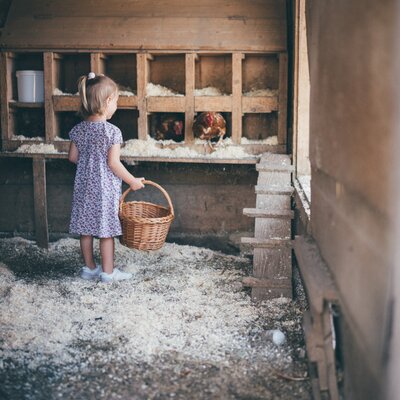 A young child in a floral dress holding a wicker basket stands in an old, rustic wooden structure filled with various items. | © Urlaub am Bauernhof / Daniel Gollner
