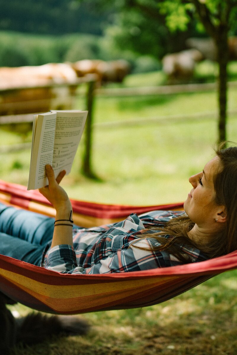 A person relaxing in a colorful hammock, reading a book in a scenic outdoor setting with trees and greenery. | © Urlaub am Bauernhof / Daniel Gollner 