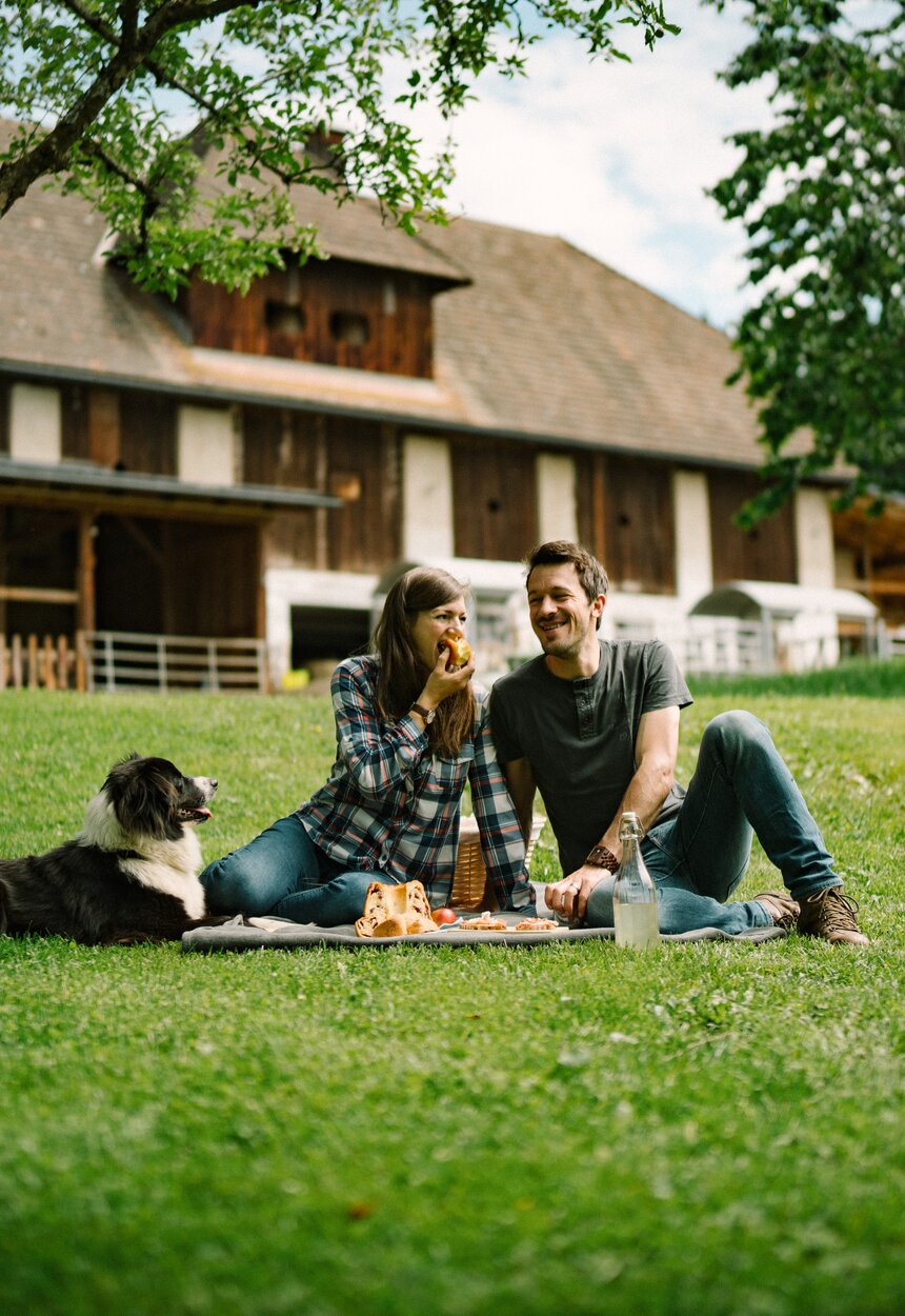 Couple relaxing on picnic blanket in front of rustic wooden building, with their dog nearby, in a lush green meadow surrounded by trees. | © Urlaub am Bauernhof / Daniel Gollner