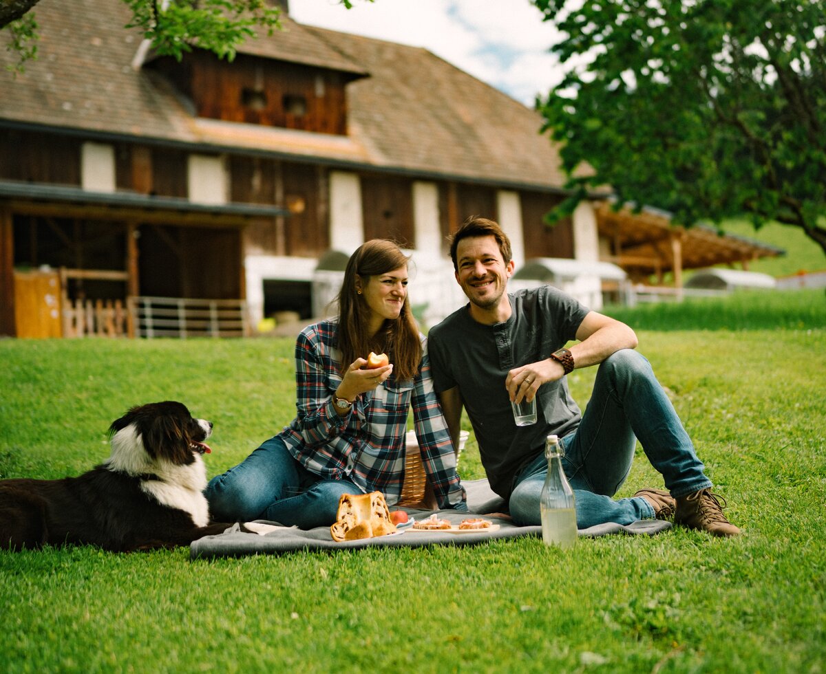 A couple relaxing outdoors with their dog, enjoying a picnic near a wooden cabin surrounded by lush greenery. | © Urlaub am Bauernhof / Daniel Gollner 
