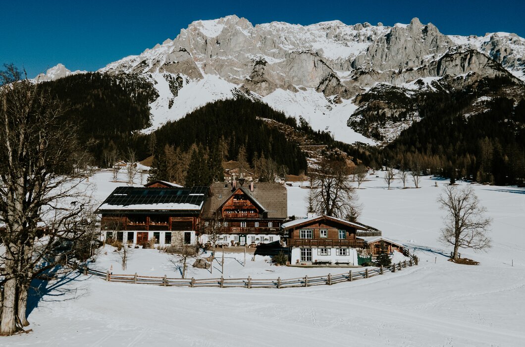 Außenansicht Hof | © Urlaub am Bauernhof / Daniel Gollner Picturesque Alpine chalets nestled in a snowy valley, framed by majestic snow-capped mountains under a clear blue sky. | © Urlaub am Bauernhof / Daniel Gollner