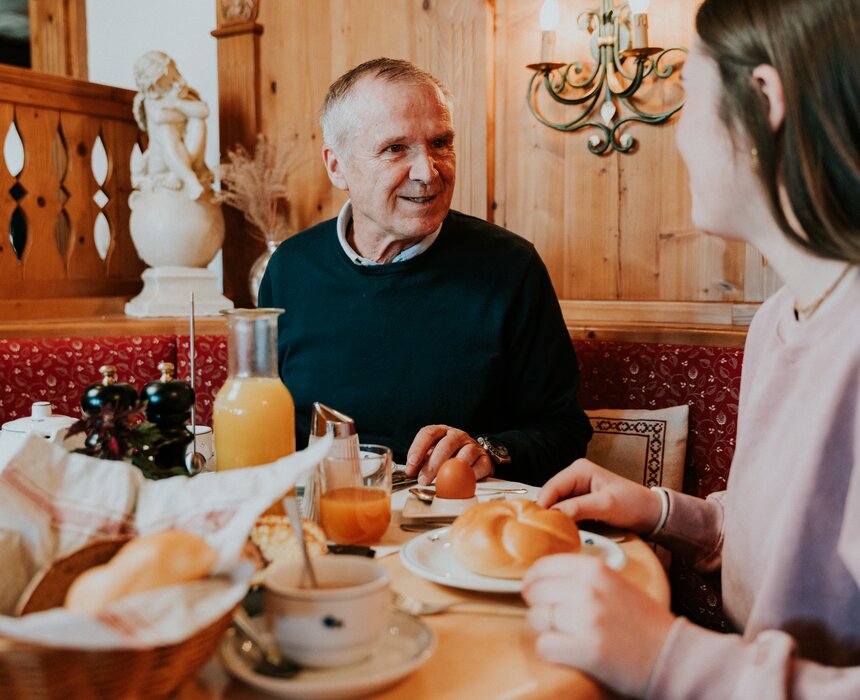 An elderly man and a young woman enjoying breakfast together in a cozy, warmly decorated room.