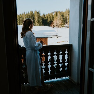 Woman in a bathrobe standing on the balcony, looking at snowy landscape with trees.