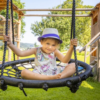 A young girl in a colorful dress and hat playing on a swing in a grassy outdoor area surrounded by a wooden structure.