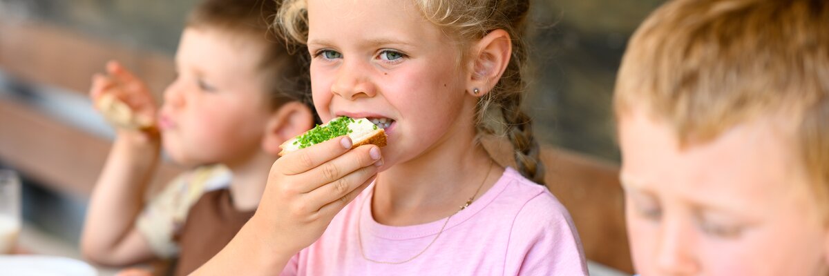 A young girl with curly blonde hair eating a sandwich at a table, seated with other children. | © Urlaub am Bauernhof / Punkt und Komma