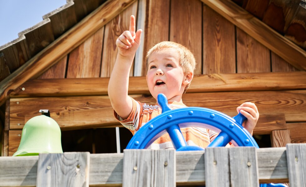 Young child playing excitedly with steering wheel toy on wooden playground structure. | © Urlaub am Bauernhof / Punkt und Komma