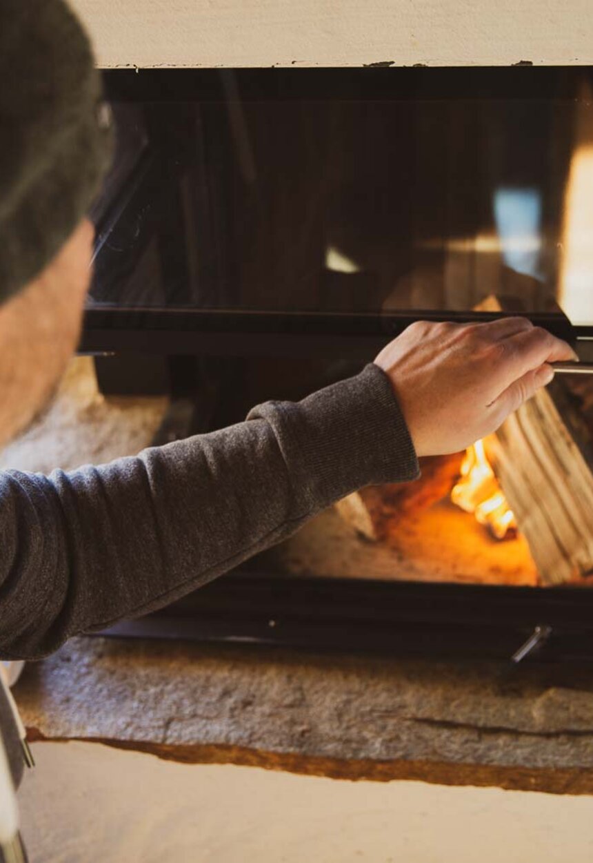 Person adding firewood to a cozy fireplace in a rustic, wooden interior. | © Urlaub am Bauernhof / Pascal Baronit