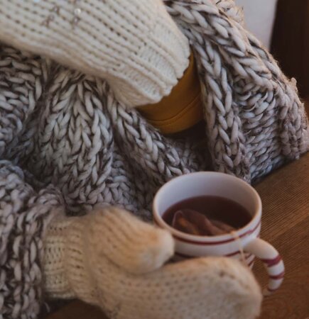 Cozy knit blanket, warm mug of tea, wooden table, close-up of comfy indoor setting. | © Urlaub am Bauernhof / Pascal Baronit