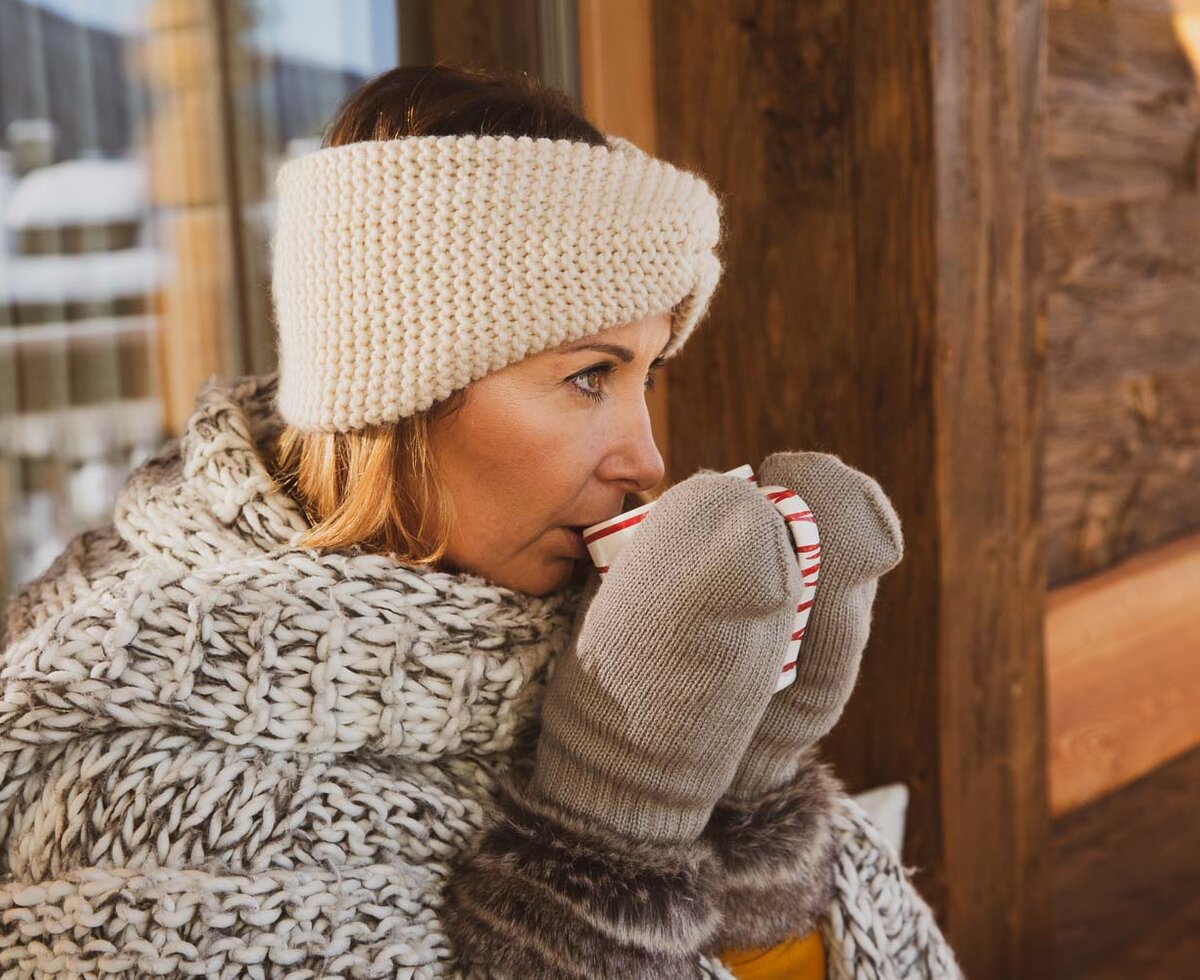 A woman in warm winter clothing, including a knitted headband and mittens, against a wooden background, appears to be taking a break or enjoying the scenery. | © Urlaub am Bauernhof / Pascal Baronit
