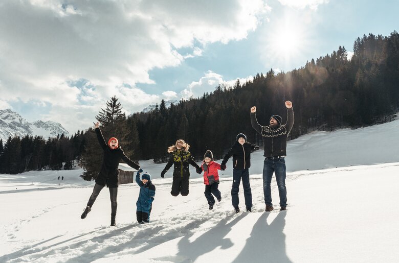 A group of people enjoying the snowy landscape, with mountains and forest in the background. They appear to be having fun and playing in the snow. | © Urlaub am Bauernhof / Punkt und Komma