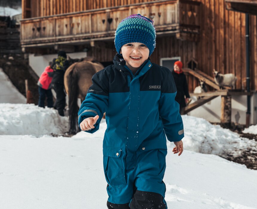 Kind im Schnee | © Urlaub am Bauernhof / Punkt und Komma A young child in a warm winter outfit, smiling and standing in a snowy mountain scene with rustic wooden buildings in the background. | © Urlaub am Bauernhof / Punkt und Komma