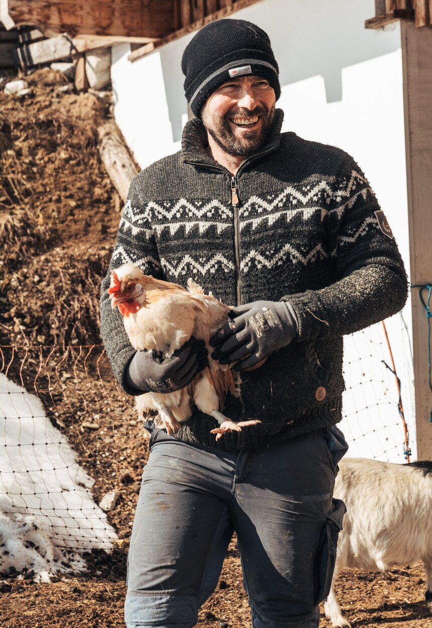 Mann mit Huhn | © Urlaub am Bauernhof / Punkt und Komma A man wearing a warm patterned jacket and hat holds a chicken while surrounded by a rural, rustic setting with an old wooden barn. | © Urlaub am Bauernhof / Punkt und Komma