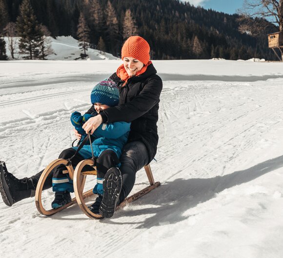 Schlittenfahrt | © Urlaub am Bauernhof / Punkt und Komma A smiling person in winter clothing sledding on a snowy landscape with a wooded mountain backdrop. | © Urlaub am Bauernhof / Punkt und Komma