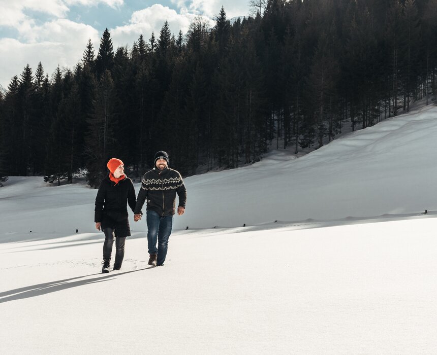 Two people walking together in a snowy winter landscape with a forested mountainous background. | © Urlaub am Bauernhof / Punkt und Komma